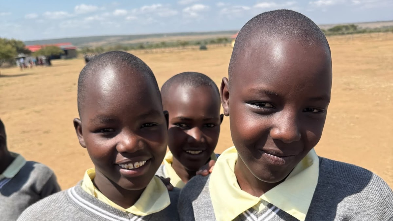 Three smiling Kenyan schoolgirls in grey uniform sweaters standing together outside their school