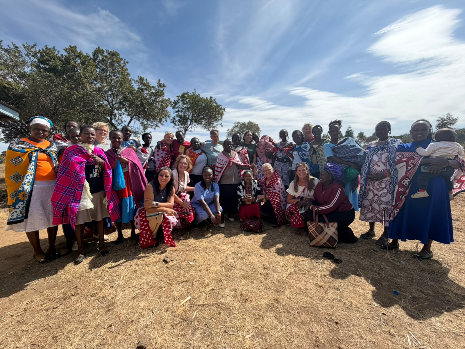 A large group of Kenyan women and a visiting team standing together in a sunlit field, many wearing brightly colored shukas and wraps
